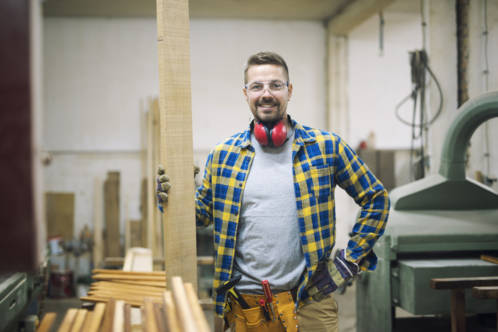 Portrait of professional middle aged carpenter with wood plank and tools standing in his woodworking workshop while workers working in background.