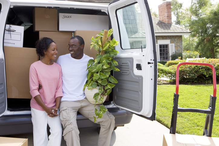Smiling couple moving house in rear of van with pot plant at camera