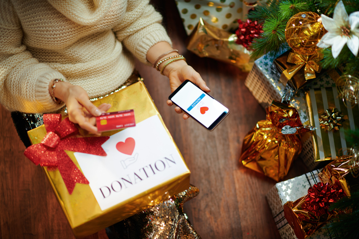 Stylish housewife in gold sequin skirt and white sweater with credit card making donation via smartphone application under decorated Christmas tree near present boxes.